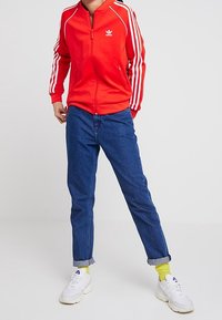 Červená bunda na zip s bílými pruhy, doplněná o modré džínové kalhoty s vyhrnutými nohavicemi. Boty jsou bílé; jsou vidět jasně žluté ponožky.