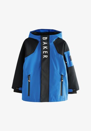 Uśmiechnięty chłopiec w niebiesko-czarnej kurtce z kapturem z napisem "Baker by Ted Baker" na zakładce zamka błyskawicznego na jednolitym szarym tle.