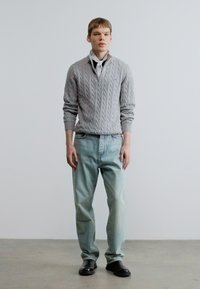 Γκρι πουλόβερ με πλέξη καλώδιο και γιακά, συνδυασμένο με ανοιχτό μπλε τζιν με ίσιο πόδι και μαύρα παπούτσια. Το φόντο είναι ουδέτερο και μίνιμαλ.