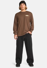 Camicia marrone a maniche lunghe con logo sul petto, abbinata a pantaloni neri a gamba larga e scarpe nere, indossata con un berretto di maglia nera.