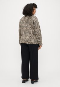 Zebra-mønsteret jakke med lange ærmer, i beige og brune nuancer, parret med brede sorte bukser og sko med blokhæle. Minimalistisk stil.