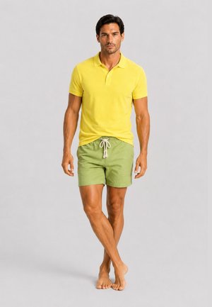 Mann steht barfuß, trägt ein gelbes Polohemd und grüne Shorts mit Kordelzug vor einem schlichten hellgrauen Hintergrund.