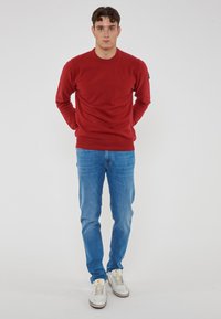 Giovane uomo in piedi con le mani dietro la schiena, che indossa un maglione rosso, jeans blu e sneaker bianche su uno sfondo chiaro.