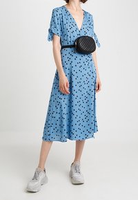 Robe bleu clair à manches courtes, imprimé floral noir, et décolleté en V. Portée avec un sac bandoulière noir et des baskets grises.