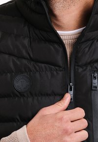 Veste matelassée noire avec capuche, arborant un logo distinctif, fermeture éclair devant et tissu texturé avec des coutures horizontales.