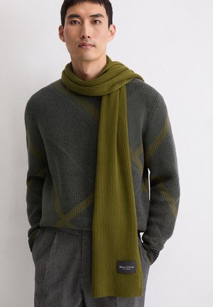 Homme portant un pull vert à motifs, une écharpe côtelée vert olive et un pantalon gris, debout contre un mur blanc uni.