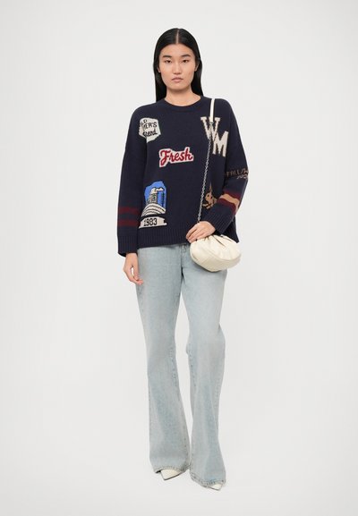 Navy knitted sweater with colorful patches, round neckline, and ribbed cuffs. Paired with light denim flared jeans and a white shoulder bag.
