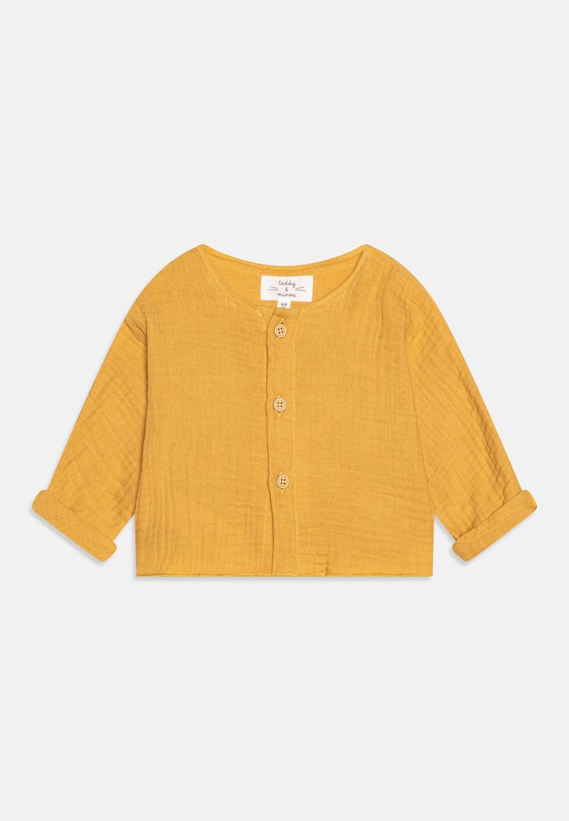 Top long jusqu'au poignet, jaune moutarde, en tissu texturé avec un col rond et trois boutons en bois sur le devant.