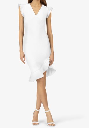 Woman wearing a white knee-length dress with ruffled sleeves and hem, paired with white ankle-strap high-heeled sandals.