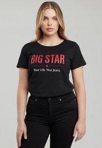 Hosszú szőke hajú nő fekete "BIG STAR Your Life. Your Jeans." feliratú pólóban és fekete farmert visel, miközben szürke háttér előtt áll.
