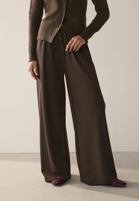 Weite braune Hosen aus weichem Material mit einem Kordelzugbund. Kombiniert mit einer taillierten braunen Strickjacke und dunkelvioletten Schuhen.