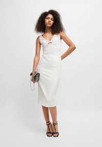 Robe blanche ajustée avec un décolleté en V et un détail découpé, fabriquée en tissu lisse. Le modèle tient une pochette noire texturée et porte des talons avec des lanières.