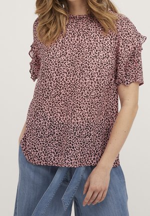 Vrouw draagt een roze blouse met luipaardprint, gerimpelde korte mouwen en blauwe jeans met wijde pijpen en een stoffen riem.