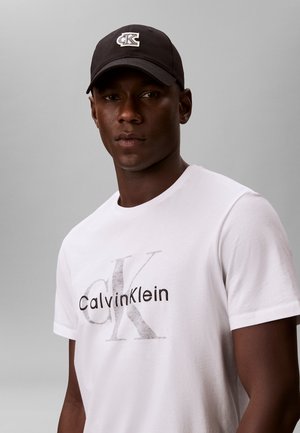 Homme portant une casquette noire avec un logo blanc et un t-shirt blanc Calvin Klein sur fond gris.