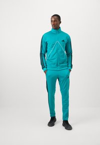Man standing with hands in pockets wearing a teal zip-up track jacket and matching pants with black stripes and black sneakers.