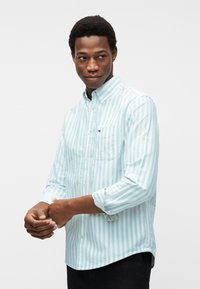 Άνδρας που ρυθμίζει τα μανίκια ενός πουκαμίσου με ανοιχτό μπλε και λευκές ρίγες και τσέπη στο στήθος, στέκεται μπροστά σε απλό λευκό φόντο.