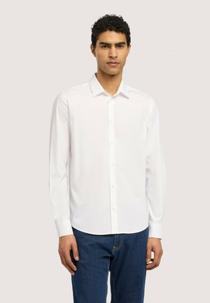 Mann trägt ein schlichtes weißes Hemd mit langen Ärmeln und blaue Jeans, steht vor einem neutralen hellen Hintergrund.