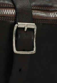 Detalle de un bolso de cuero marrón que presenta un hebilla de metal plateado y un cierre de cremallera con dientes visibles, mostrando una superficie texturizada y rica.