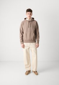 Bruine hoodie met een voorkraagzak, crèmekleurige, losse broek en lichtbruine schoenen tegen een neutrale achtergrond.