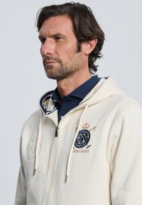 Sudadera con capucha cremalleras crema con tela texturizada, logo bordado con corona y mazos cruzados, y cordones. Camiseta tipo polo azul marino debajo.