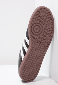 Suola di sneaker nera Adidas con grip texturizzato marrone, tre strisce bianche sul lato, esposta contro uno sfondo angolare bianco.