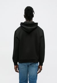 Sweatshirt preta com capuz, corte solto, punhos e cintura canelados, vista de costas sobre um fundo branco liso.