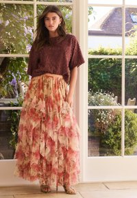 Texturierter, bordeauxroter kurzer Pullover kombiniert mit einem fließenden, gestuften Maxirock mit floralem Muster in Rosa- und Cremetönen. Goldene Riemchenabsätze.