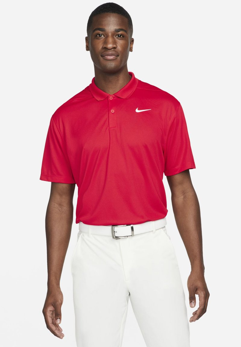 Röd polo skjorta i slät tyg med krage, prydd med en vit Nike-logotyp på vänster bröst. Kombinerad med vita byxor och ett bälte.