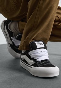 Persoană îmbrăcată în pantaloni maro și adidași Vans alb-negru, așezată pe un podea gri, arătând detaliile pantofilor și dunga de la șireturile acestora.