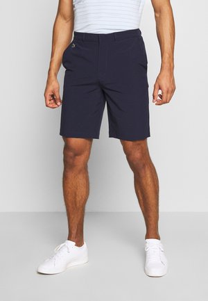 Homme portant un short bleu marine, des baskets blanches et une chemise rayée bleu clair, debout devant un fond uni.
