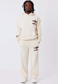 Crème hoodie en bijpassende joggers van zachte stof, met bruine stiksels in kruisvorm en een voorzak. Witte sneakers.