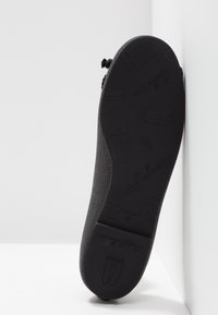 Chaussure noire plate avec une surface texturée et un bout rond. La semelle présente des motifs en relief, y compris le nom de la marque et des motifs.