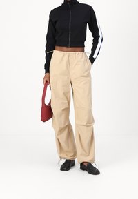 Svart kort zip-up sweatshirt, beiga byxor med dragsko, svarta loafers och en liten röd handväska. Släta texturer, avslappnad passform, minimal detaljering.