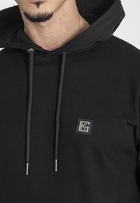 Felpa nera con cappuccio realizzata in tessuto morbido, con cordini grigi, una tasca a marsupio e un'etichetta quadrata sul lato sinistro del petto.