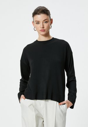 Schwarzer Pullover mit langen Ärmeln, geripptem Kragen und Saum, im entspannten Schnitt und mit glatter Textur. Kombiniert mit weißen Nadelstreifen-Hosen.