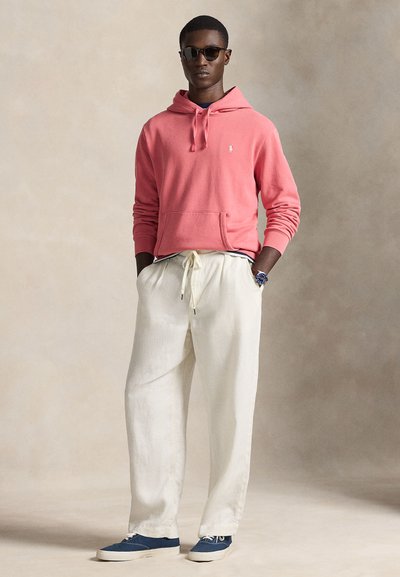 Κόκκινο φούτερ με κουκούλα και μπροστινή τσέπη, παντελόνι από κρεμ λινό και μπλε καμβά παπούτσια. Αξεσουάρ περιλαμβάνουν στρογγυλά γυαλιά ηλίου και ένα ρολόι χειρός.