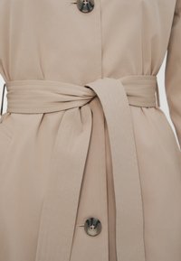 Béžový trenčkot s vázaným látkovým opaskem a tmavými kulatými knoflíky na přední straně, které zdůrazňují oblast pasu.