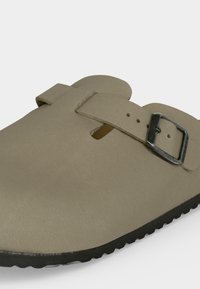 Μπεζ παπούτσι τύπου slip-on με μαύρη σόλα, που διαθέτει ένα ρυθμιζόμενο λουράκι και ασημένια αγκράφα στο πάνω μέρος.