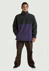 Uomo in piedi che indossa un pullover in pile a mezza zip nero e viola, pantaloni marrone scuro e scarpe slip-on nere con suole bianche.