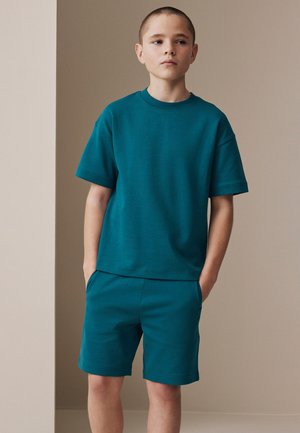 Dreng med kort hår står med hænder i lommerne iført matchende teal kortærmet skjorte og shorts mod en neutral baggrund.