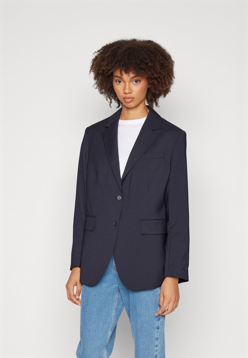 Blazer marine à la coupe structurée, avec revers crantés, deux poches avant et un fermoir à boutons, associé à un t-shirt blanc et à un jean clair.