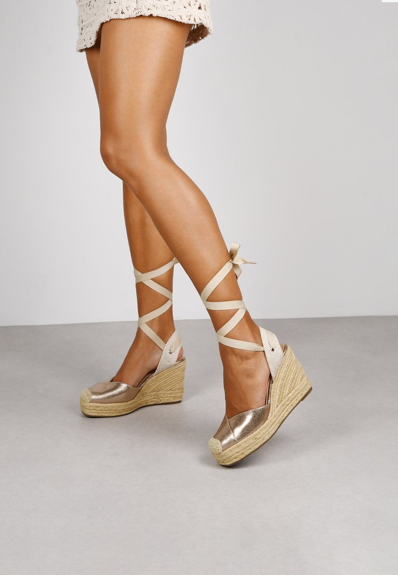 Espadrilles de cuña con parte superior de metal dorado, entresuela de yute trenzado y tiras de cinta entrecruzadas alrededor del tobillo, sobre un fondo neutro.