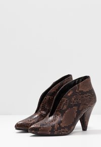 Botas de tobillo de imitación de piel de serpiente marrón con punta afilada, superficie texturizada y un tacón de 12,7 cm. Presentan un diseño con corte lateral abierto.