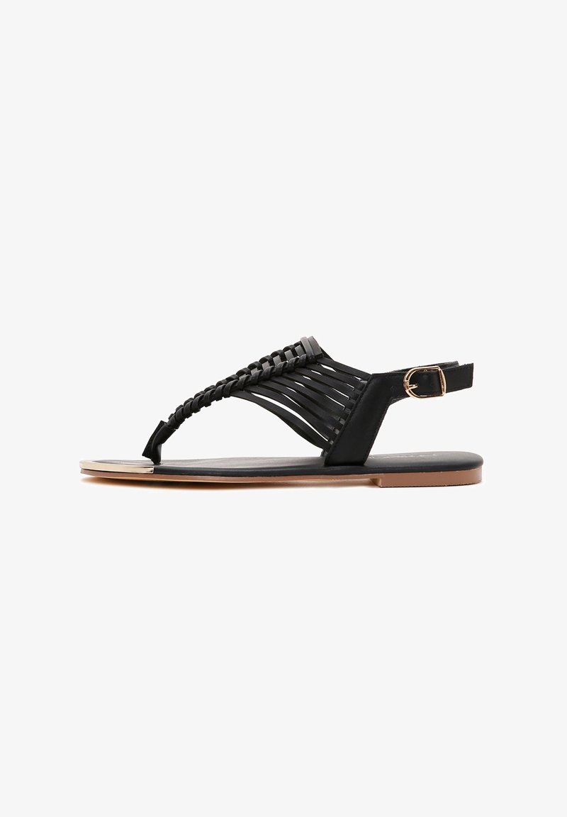 Black sandal featuring braided straps, an open toe design, a side buckle closure, and a flat sole with a metallic accent on the toe.