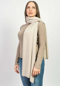 Beige tørklæde lagt om en persons hals, lavet af et blødt materiale med en glat tekstur, der komplementerer en lysbrun sweater og denim jeans.