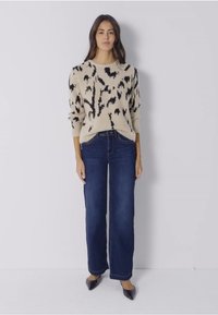 Beżowy sweter z czarnym abstrakcyjnym wzorem, połączony z ciemnoniebieskimi spodniami typu wide-leg. Stylizacja charakteryzuje się luźnym krojem i prążkowanymi mankietami.