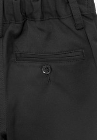 Zwarte broek met een gestructureerde stof, elastische tailleband en een achterzak met een zwarte knoopsluiting. Eenvoudig, functioneel ontwerp.