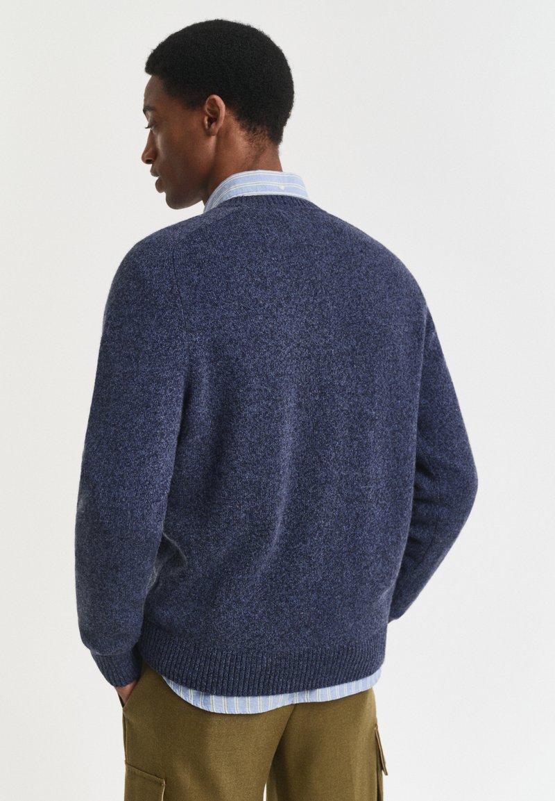 Uomo che indossa un maglione blu con scollo rotondo sopra una camicia a righe blu e pantaloni cargo color oliva, in piedi su uno sfondo bianco semplice.