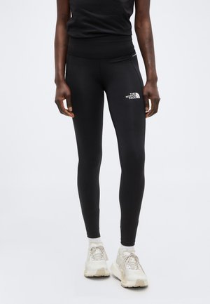 Persona con leggings negros The North Face y zapatillas deportivas blancas, de pie con los brazos relajados a los lados contra un fondo liso.