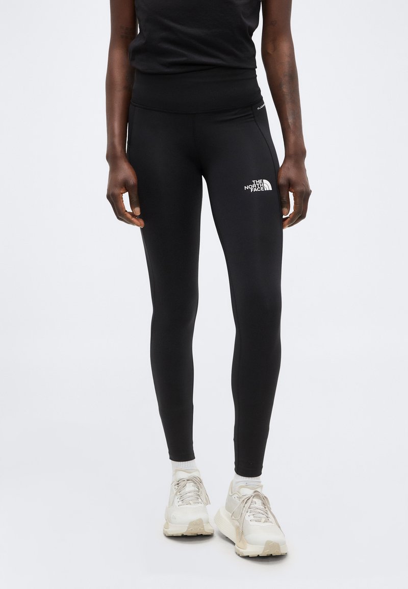 Άτομο με μαύρα κολάν The North Face και λευκά αθλητικά παπούτσια, στέκεται με τα χέρια χαλαρά στα πλευρά μπροστά σε απλό φόντο.